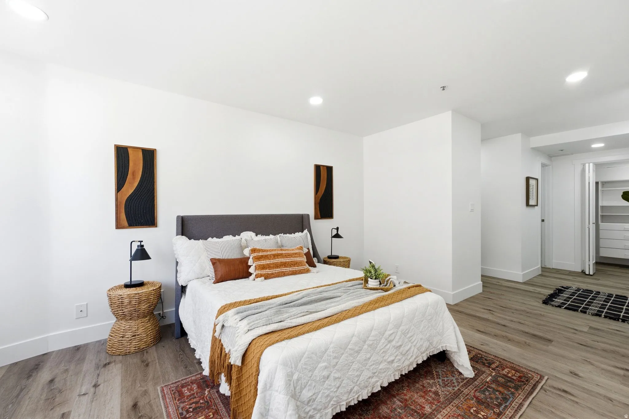 Bright and spacious master bedroom featuring light wood-finish flooring, white walls, recessed lighting, and a walk-in closet with built-in shelving and drawers
