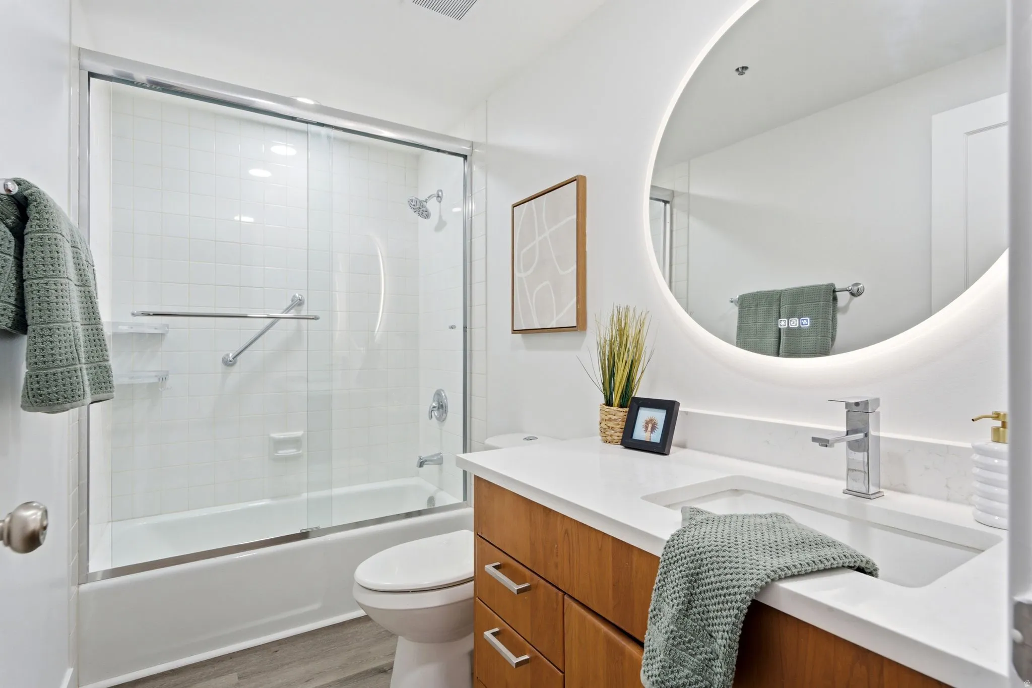 Bathroom featuring a white subway tile shower, sliding glass shower doors, a built-in bathtub, a wood-finish vanity with an integrated sink, and an illuminated circular mirror