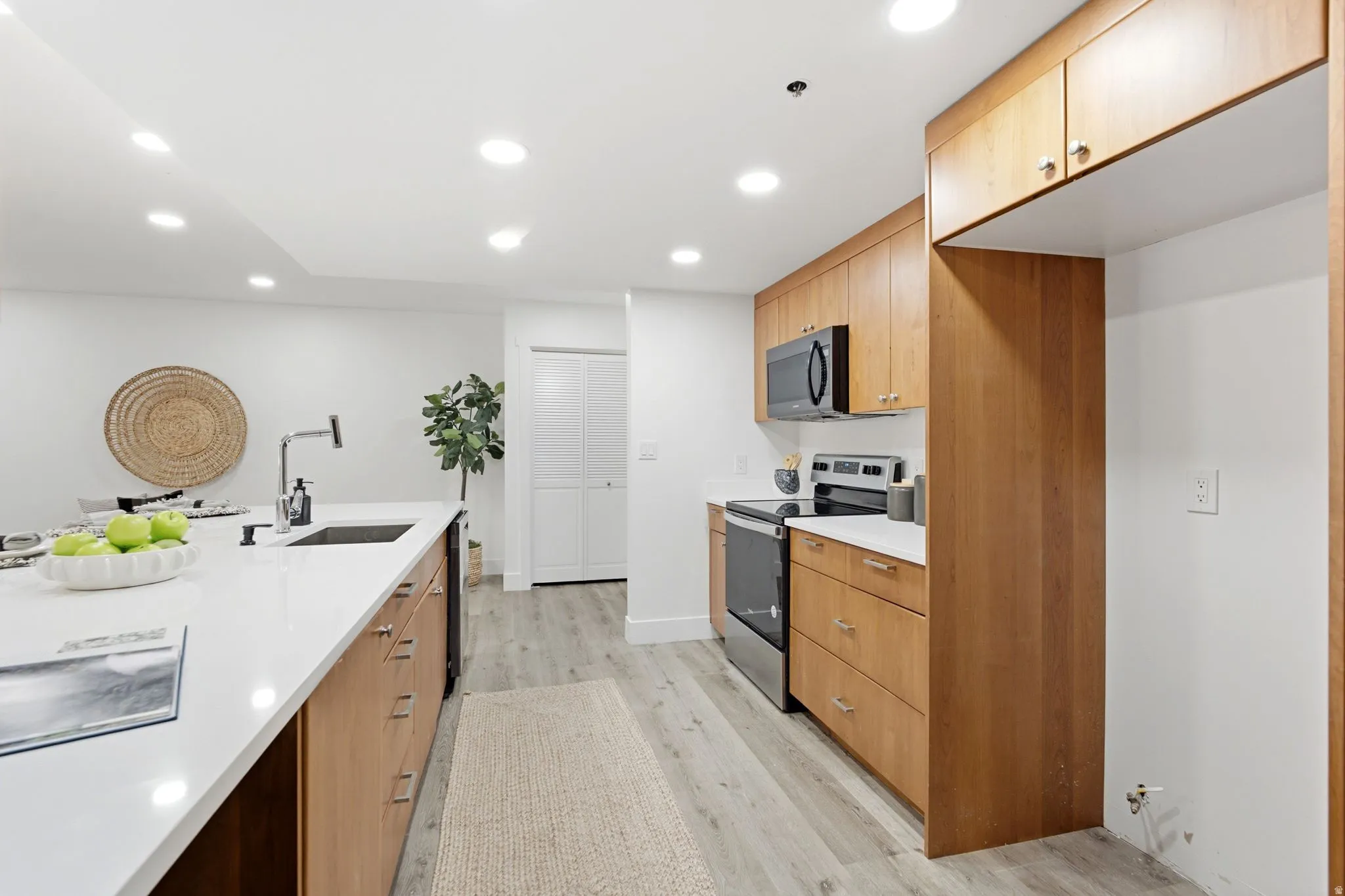 Kitchen featuring light wood-finish cabinetry, white countertops, built-in stainless-steel appliances, a kitchen island with an undermount sink and matte black faucet, and wood-finish flooring