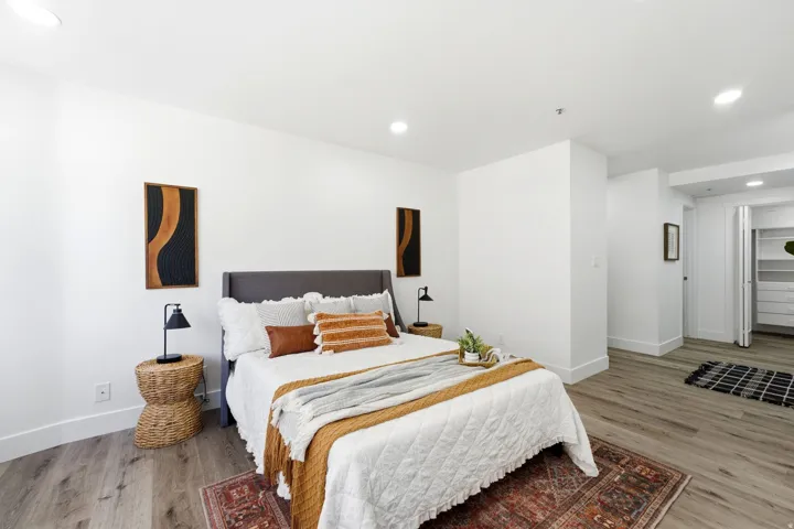 Bright and spacious master bedroom featuring light wood-finish flooring, white walls, recessed lighting, and a walk-in closet with built-in shelving and drawers