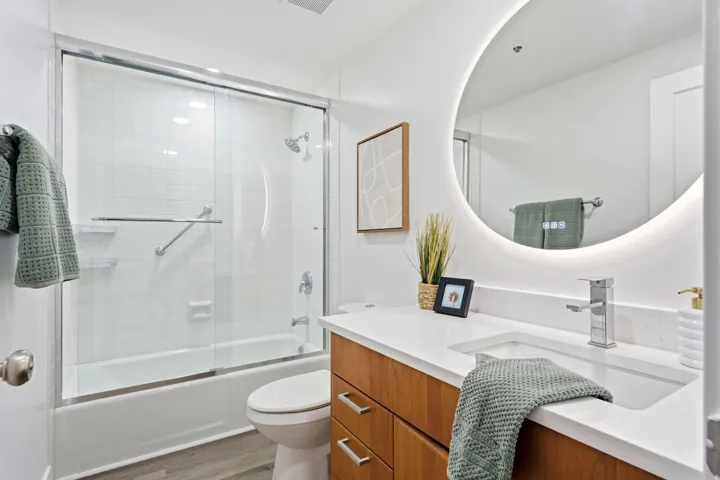 Bathroom featuring a white subway tile shower, sliding glass shower doors, a built-in bathtub, a wood-finish vanity with an integrated sink, and an illuminated circular mirror