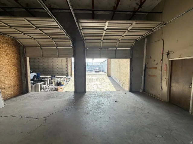 Concrete floor space with two open roll-up doors, exposed ceiling joists, and a partial plywood wall