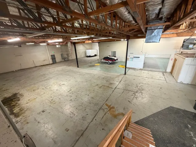 Spacious warehouse area featuring exposed wood beam ceilings, concrete flooring, and multiple fluorescent lighting fixtures