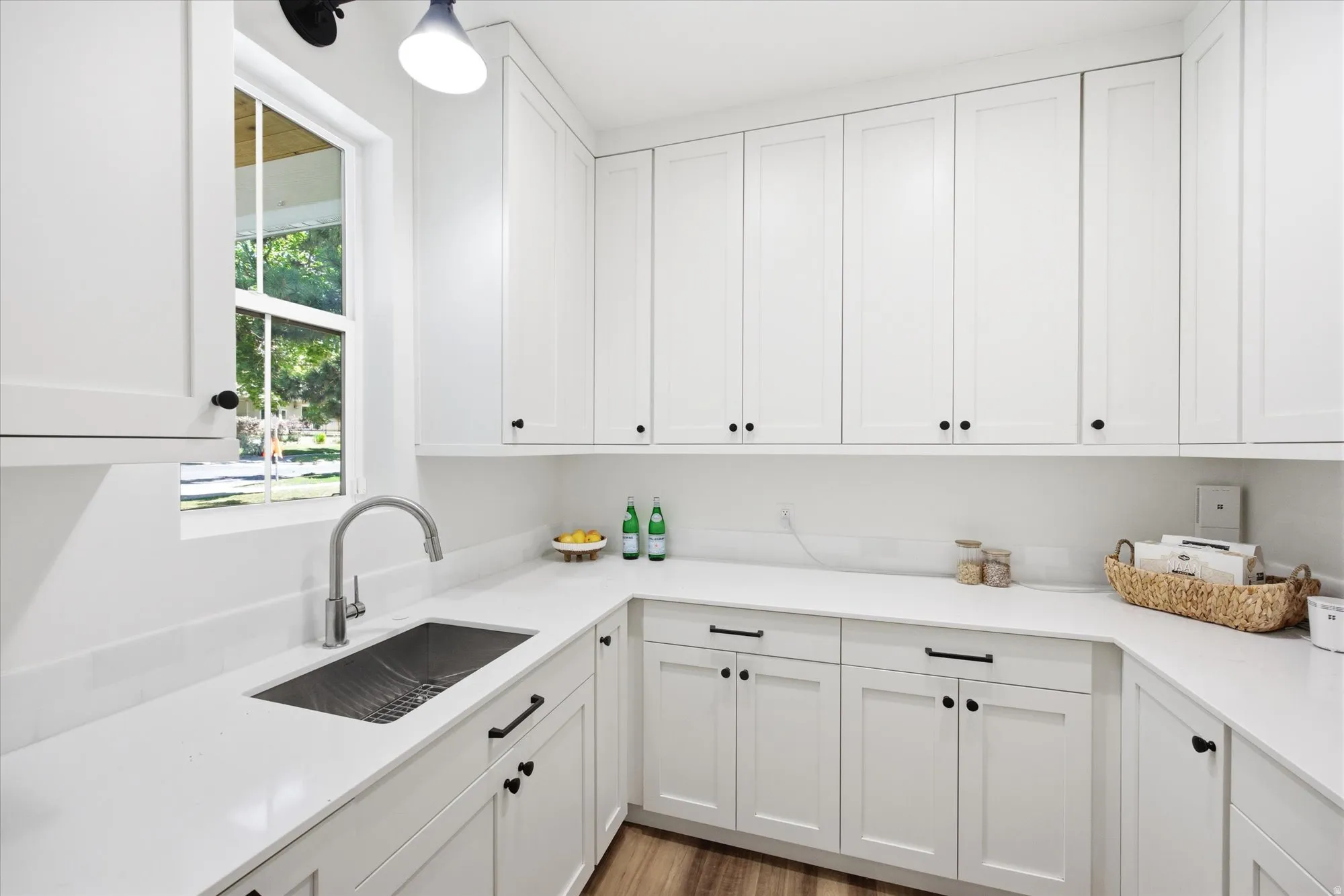 White shaker cabinetry with black hardware, white countertops, an undermount stainless steel sink with a gooseneck faucet, and wood-finish flooring