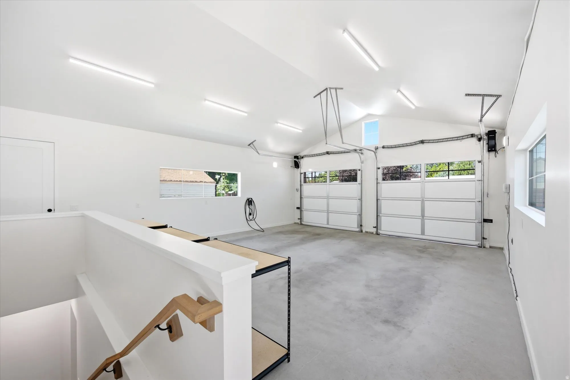 Spacious garage featuring two overhead doors with window panels, a vaulted ceiling, and multiple fluorescent light fixtures