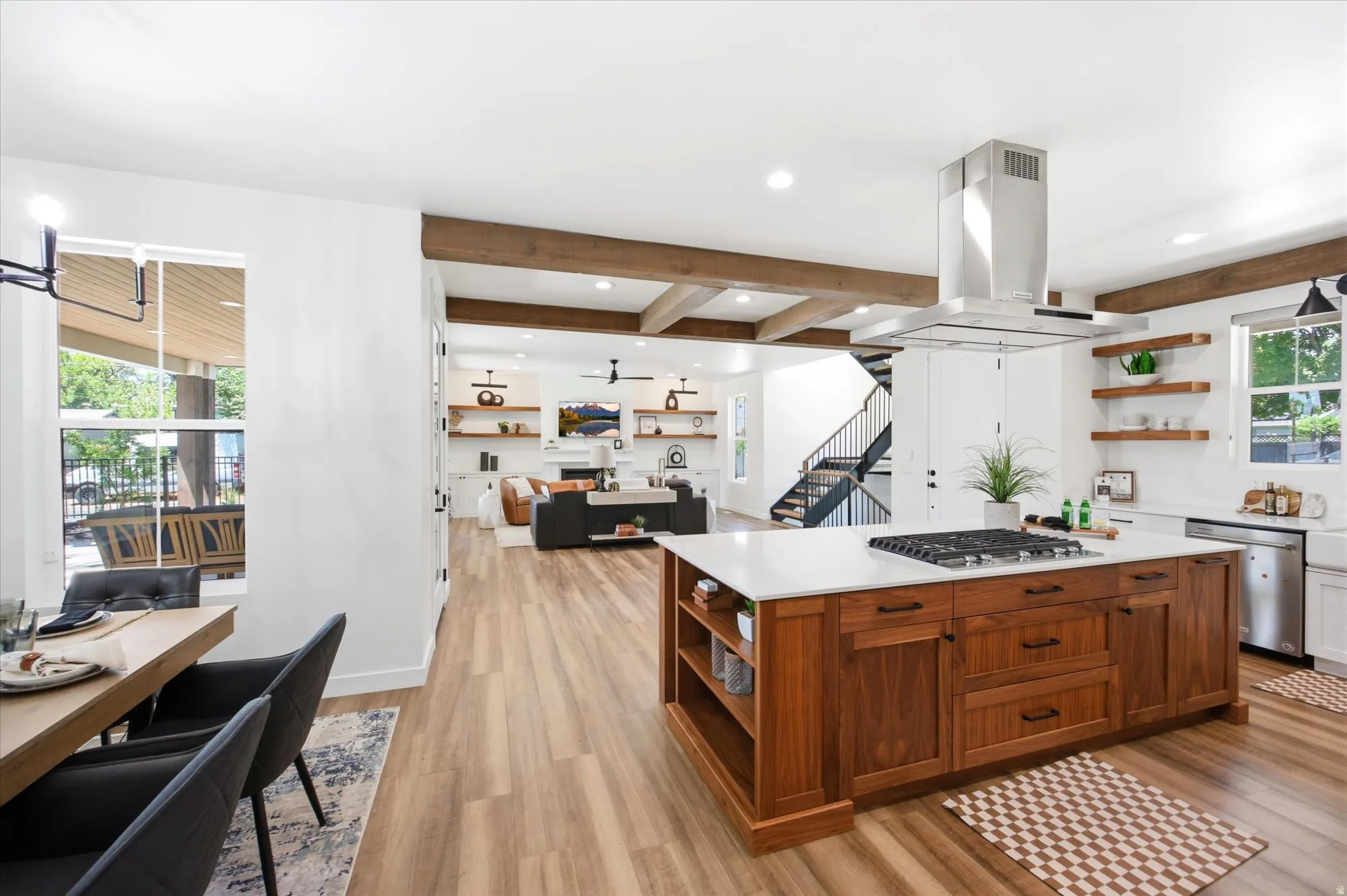 Expansive open-concept living area featuring a kitchen island with a gas cooktop, wood-finish flooring, exposed ceiling beams, a white brick fireplace, and a staircase with black metal railings
