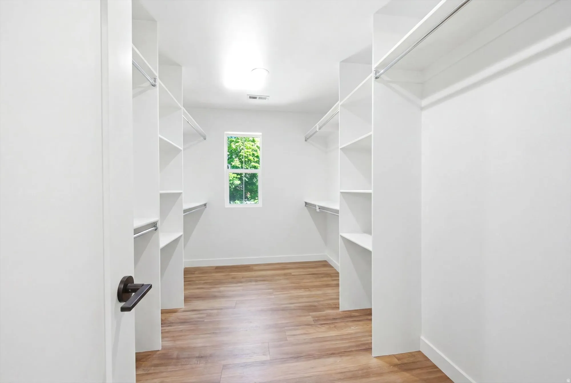 Walk-in closet featuring wood-finish flooring, white built-in shelving, hanging rods, and a window for natural illumination