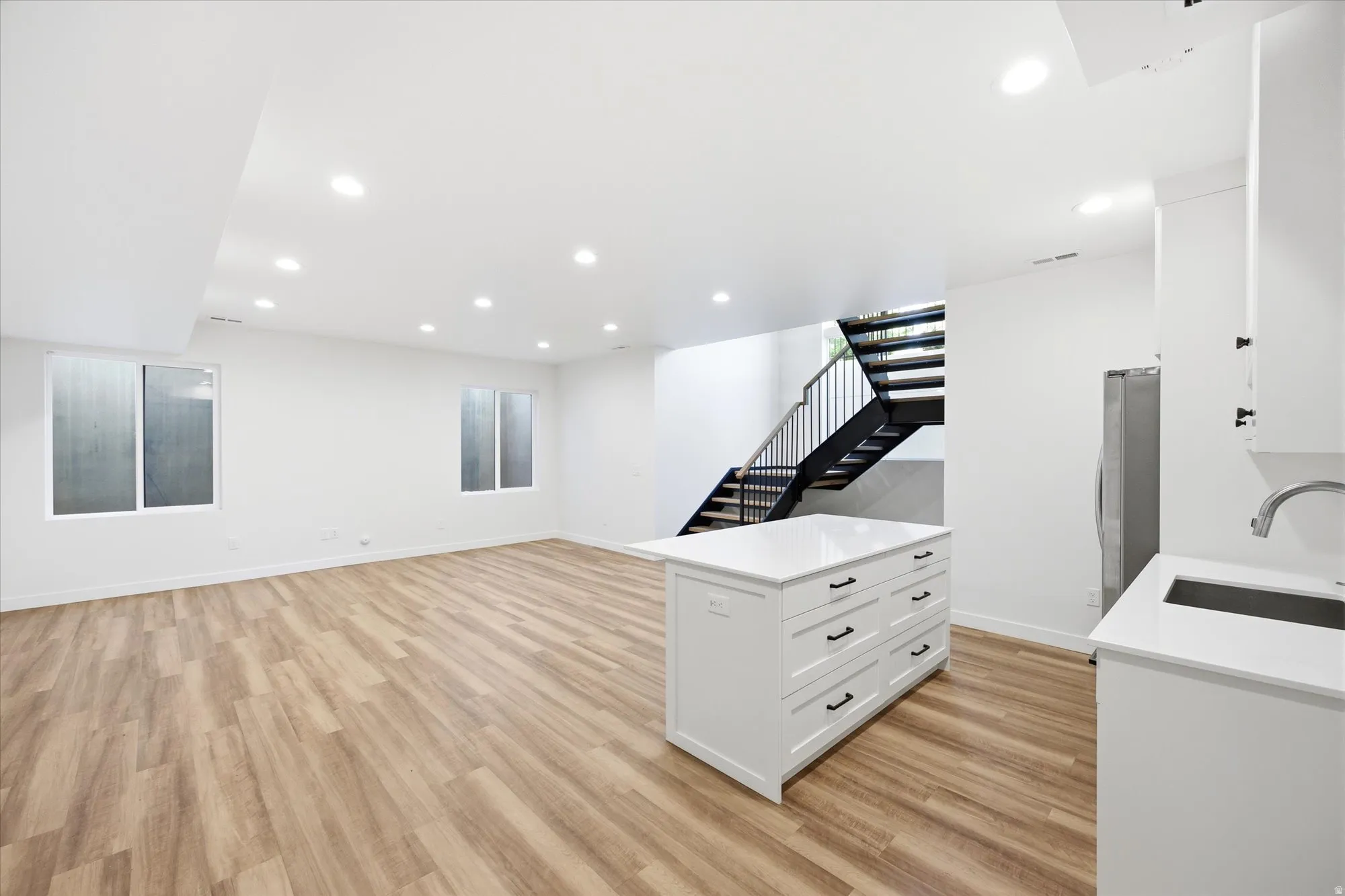 Open-concept lower level featuring recessed lighting, wood-finish flooring, a contemporary black metal staircase, white cabinetry with a sink, and a kitchen island with a white countertop