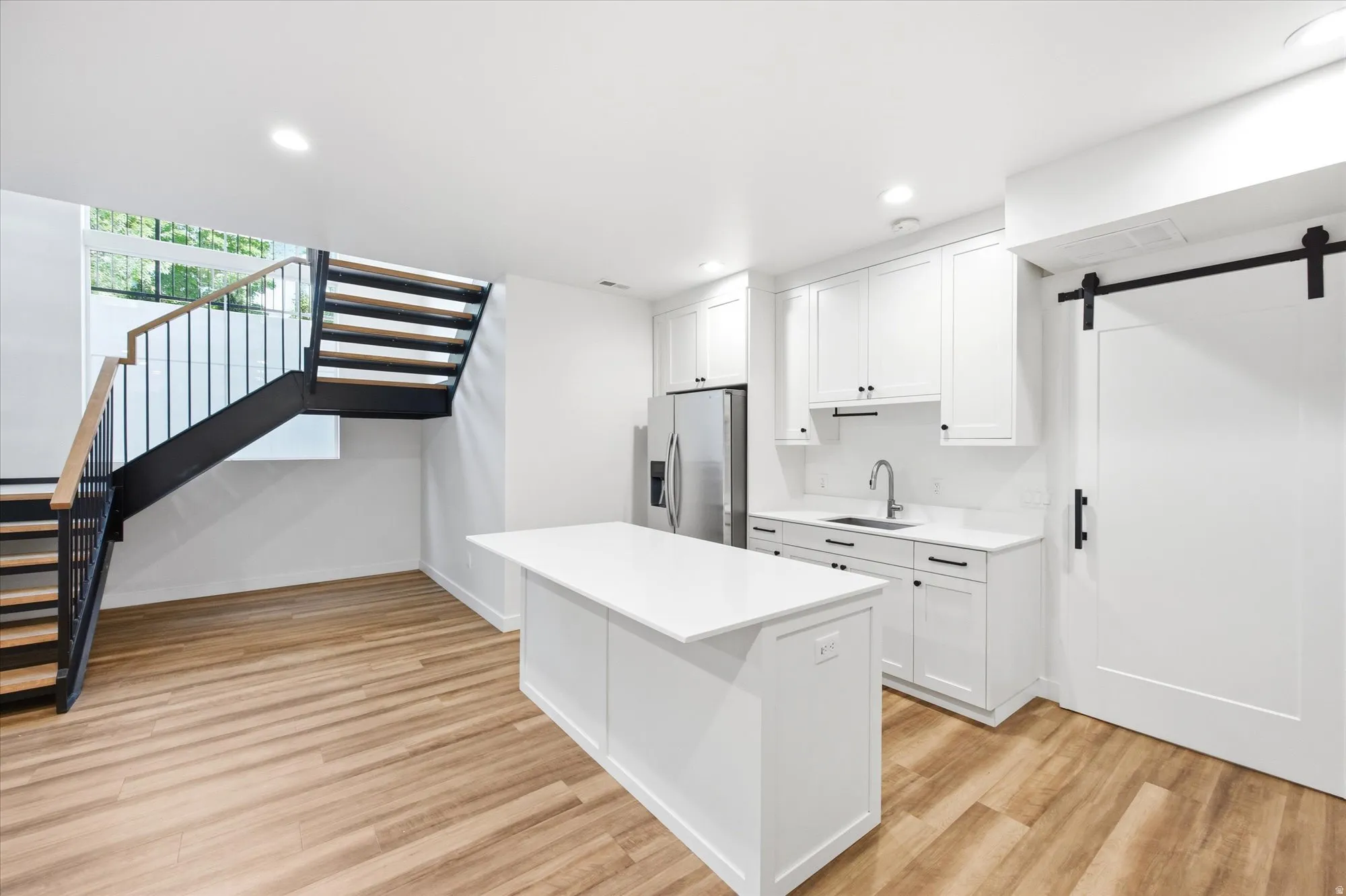 Contemporary kitchen and living area featuring a white kitchen island with quartz countertop, white shaker cabinetry, stainless steel refrigerator, and wood-finish flooring