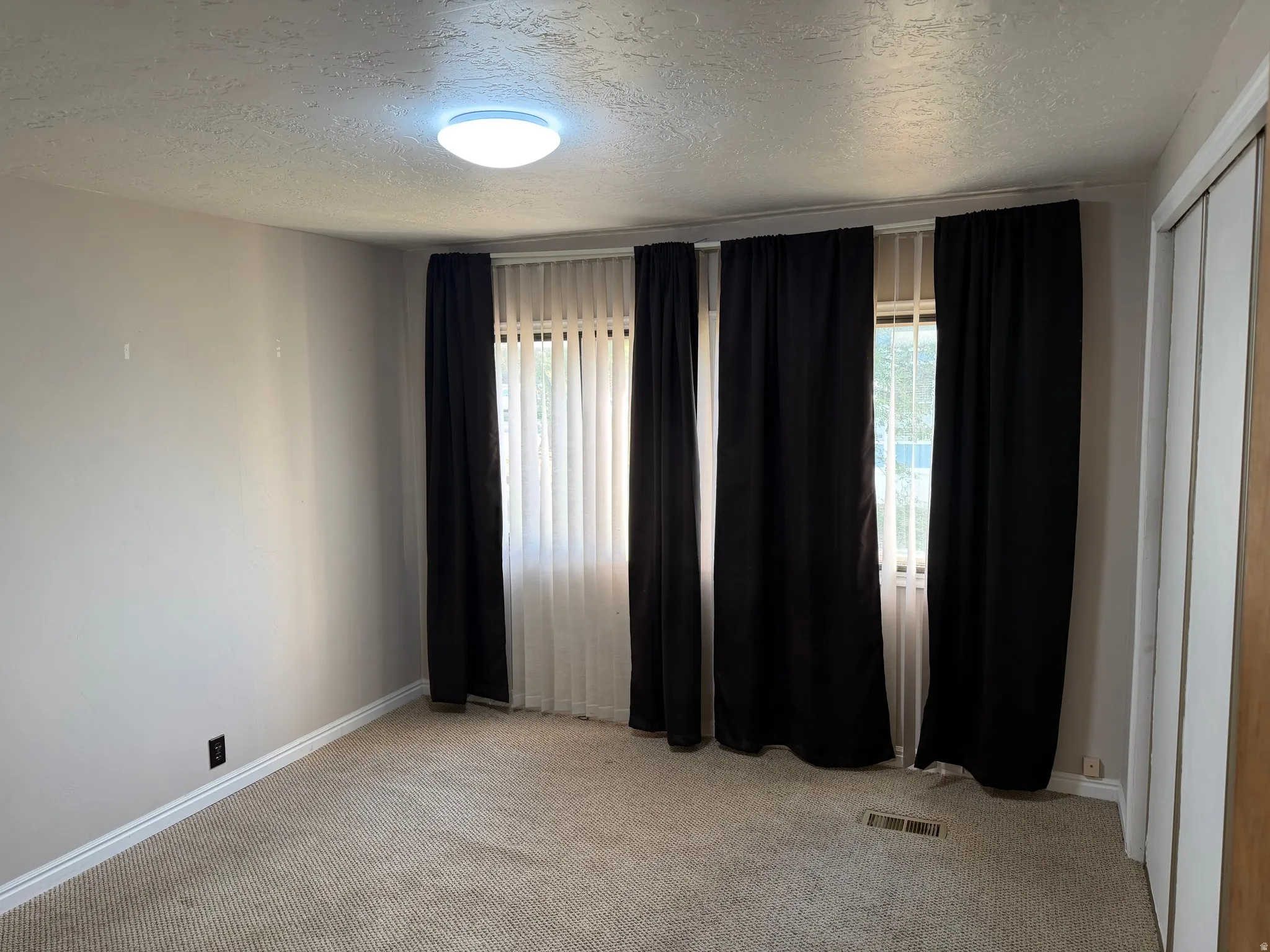 Carpeted interior space with a window featuring black and sheer white curtains, a flush-mount ceiling light fixture, and textured ceilings