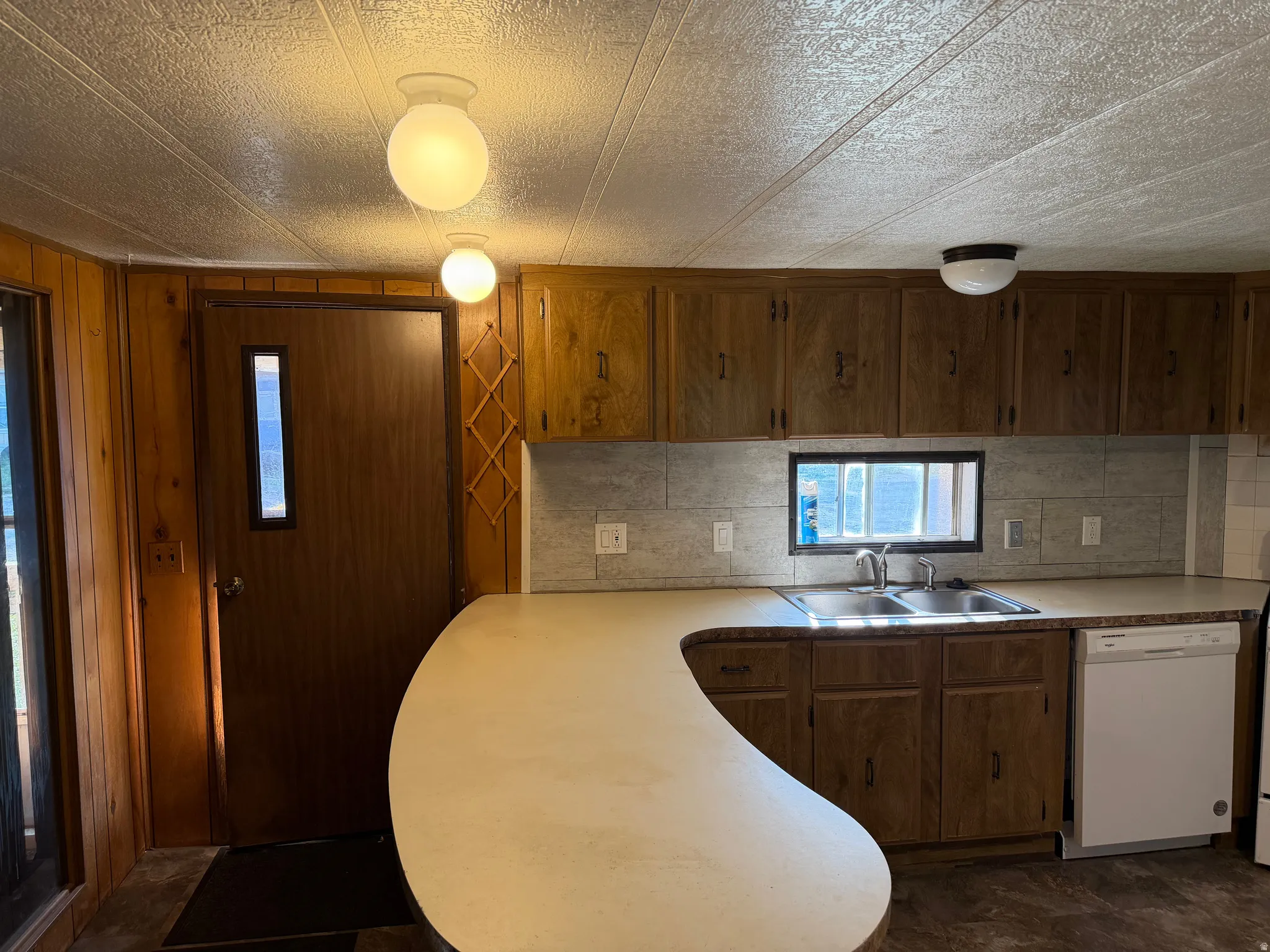Kitchen featuring a curved peninsula with a light-toned countertop, wood-finish cabinetry, dual basin sink, and a built-in dishwasher