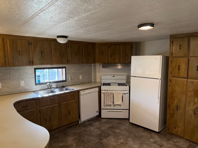 Kitchen featuring wood-finish cabinetry, solid surface countertops, a double-basin stainless steel sink, tile backsplash, and white appliances