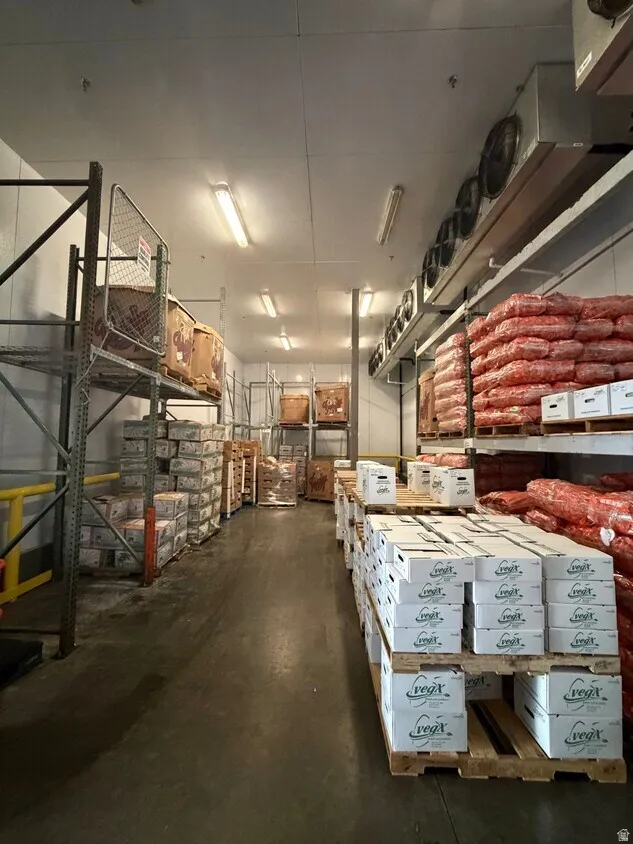 Industrial cold storage interior featuring high ceilings with fluorescent strip lighting, extensive metal racking systems, concrete flooring, and multiple wall-mounted refrigeration units