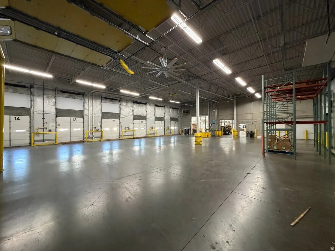 Spacious industrial warehouse featuring multiple overhead doors, high ceilings with exposed trusses, an industrial ceiling fan, and extensive fluorescent lighting