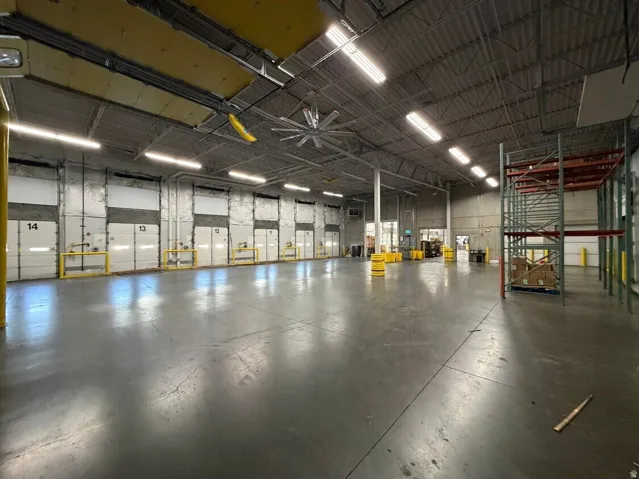 Spacious industrial warehouse featuring multiple overhead doors, high ceilings with exposed trusses, an industrial ceiling fan, and extensive fluorescent lighting