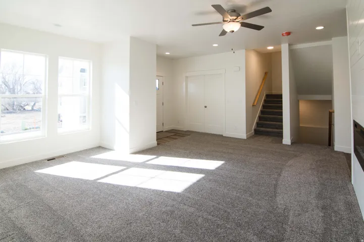 Spacious interior featuring neutral carpeting, multiple windows, a ceiling fan with integrated lighting, a built-in staircase, and a doorway