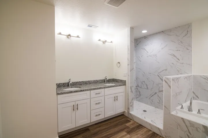 Dual vanity featuring granite countertops and undermount sinks, complemented by a walk-in shower with marble-look tile, a built-in tub surround, and wood-finish flooring