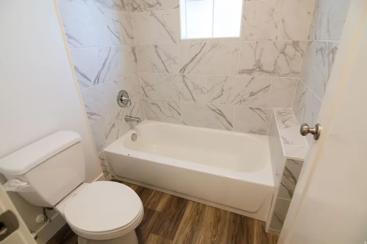 Bathroom featuring a white bathtub, chrome fixtures, white tile surround with grey veining, wood-finish flooring, and a white toilet