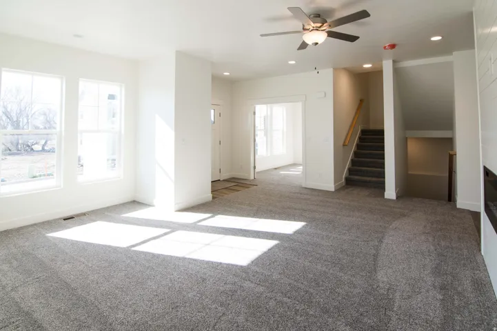 Spacious living area featuring light gray carpeting, multiple large windows, and a ceiling fan with integrated lighting