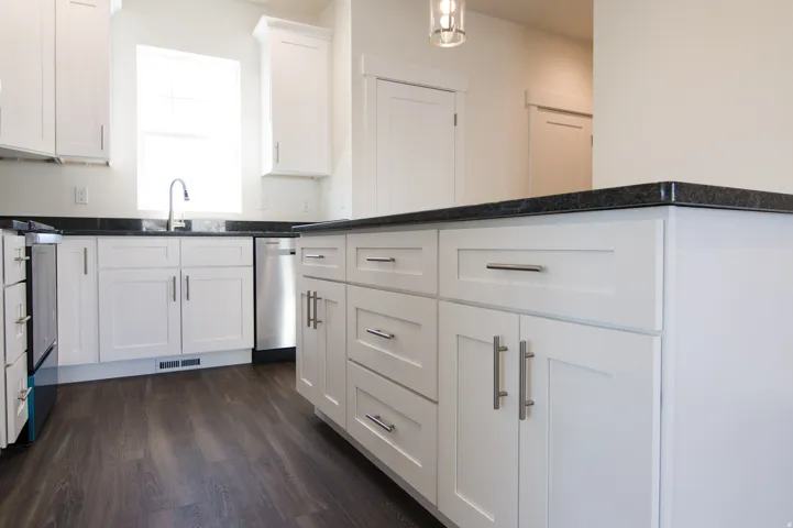 Kitchen featuring dark wood-finish flooring, white shaker cabinetry with brushed nickel hardware, dark countertops, a stainless steel dishwasher, and a gooseneck faucet