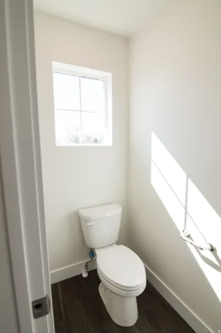 White toilet with wood-finish flooring, white baseboards, and a framed window
