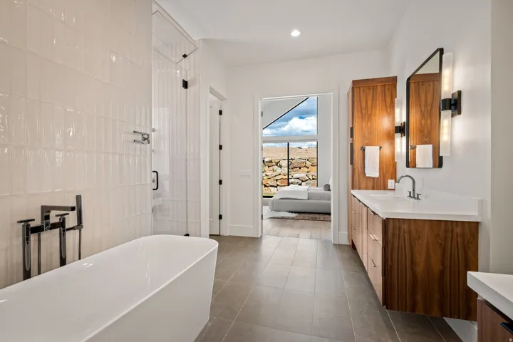 Contemporary bathroom featuring a freestanding tub with matte black fixtures, a walk-in shower with clear glass enclosure, a dual vanity with wood-finish cabinetry and white countertops, and sleek wall-mounted lighting