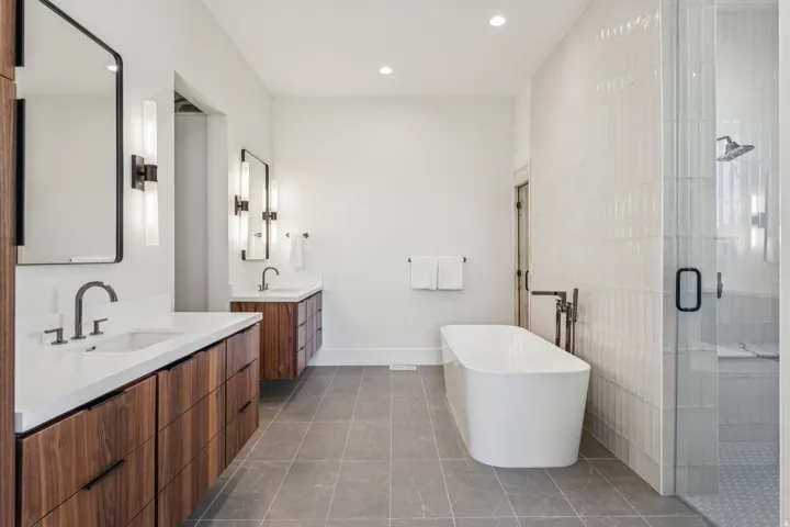 Contemporary bathroom featuring a freestanding soaking tub, a frameless glass shower enclosure with a built-in bench, dual vanities with wood-finish cabinetry, integrated sinks, and modern sconce lighting