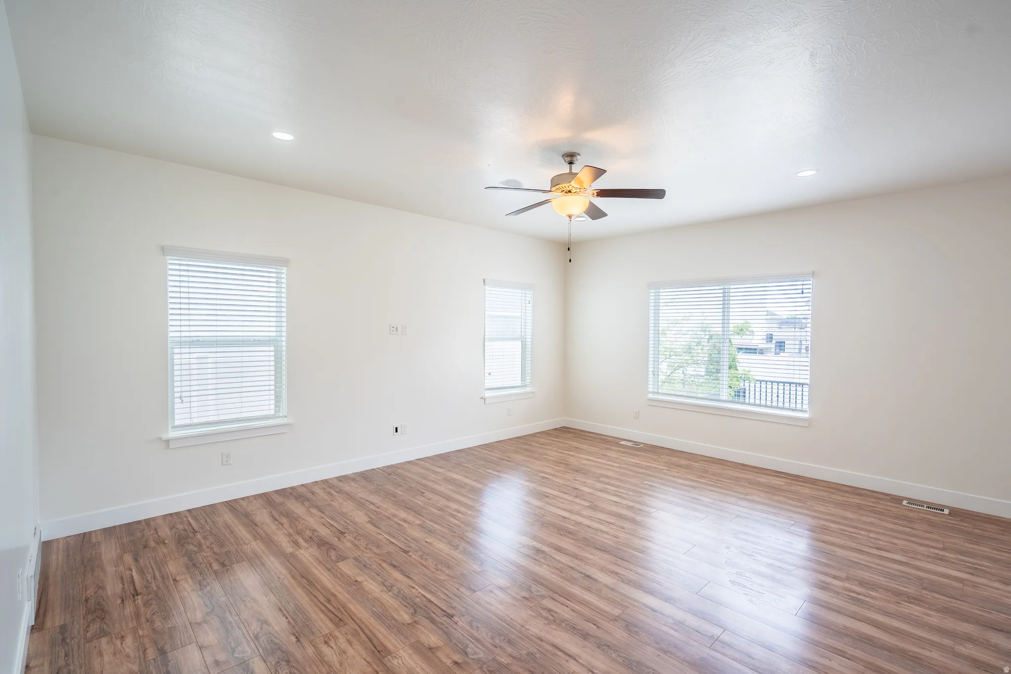 Spacious room featuring wood-finish flooring, white walls, multiple windows with blinds, a ceiling fan with light, and recessed lighting