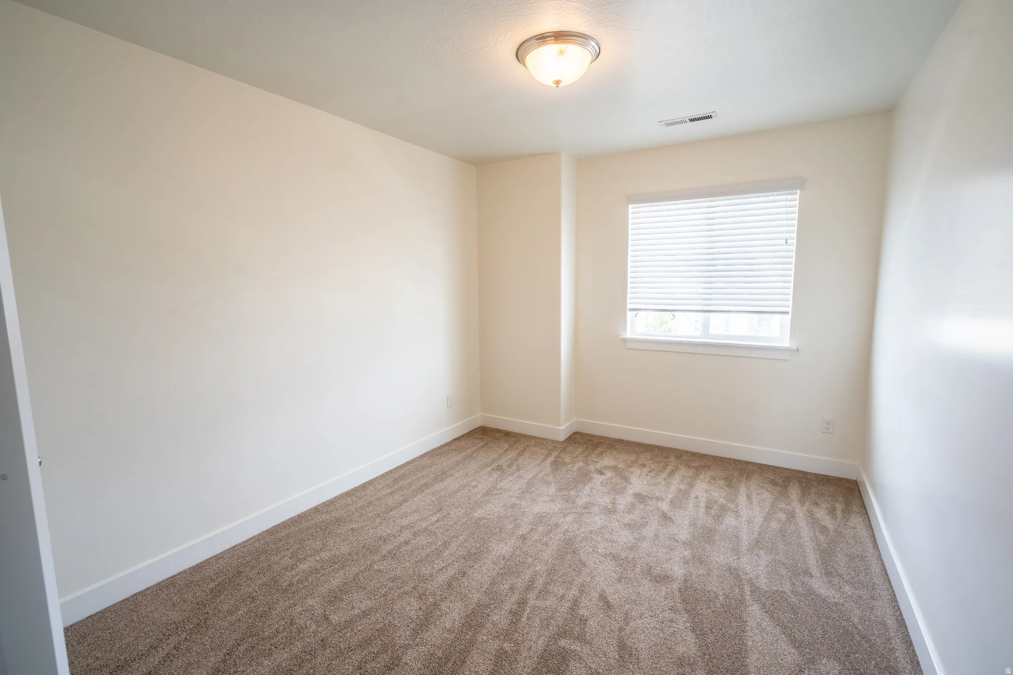 Neutral-toned room with plush carpeting, a single window featuring horizontal blinds, and a semi-flush mount ceiling light fixture