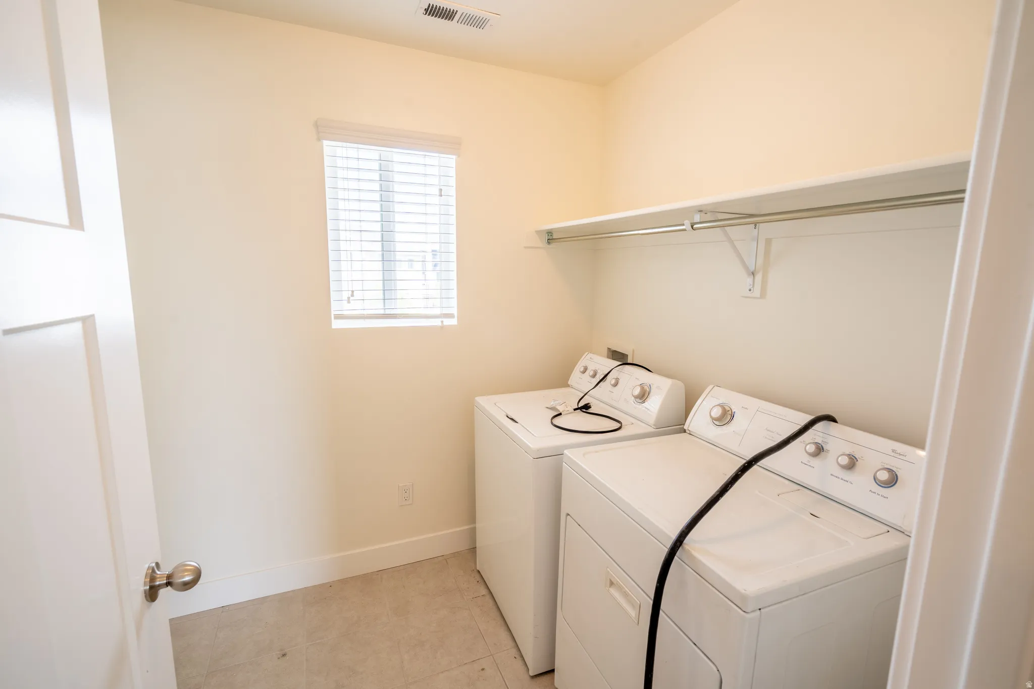 Dedicated laundry area featuring a built-in shelf and hanging rod, a window with horizontal blinds, and tile flooring