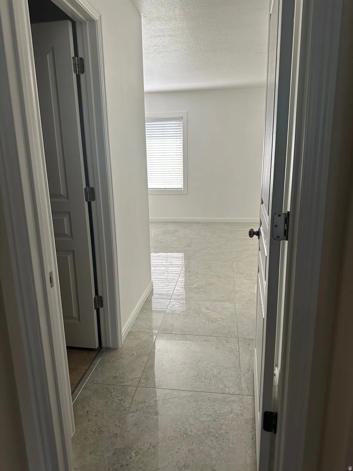 Hallway with polished tile flooring, leading to a room with a window and white louvered blinds