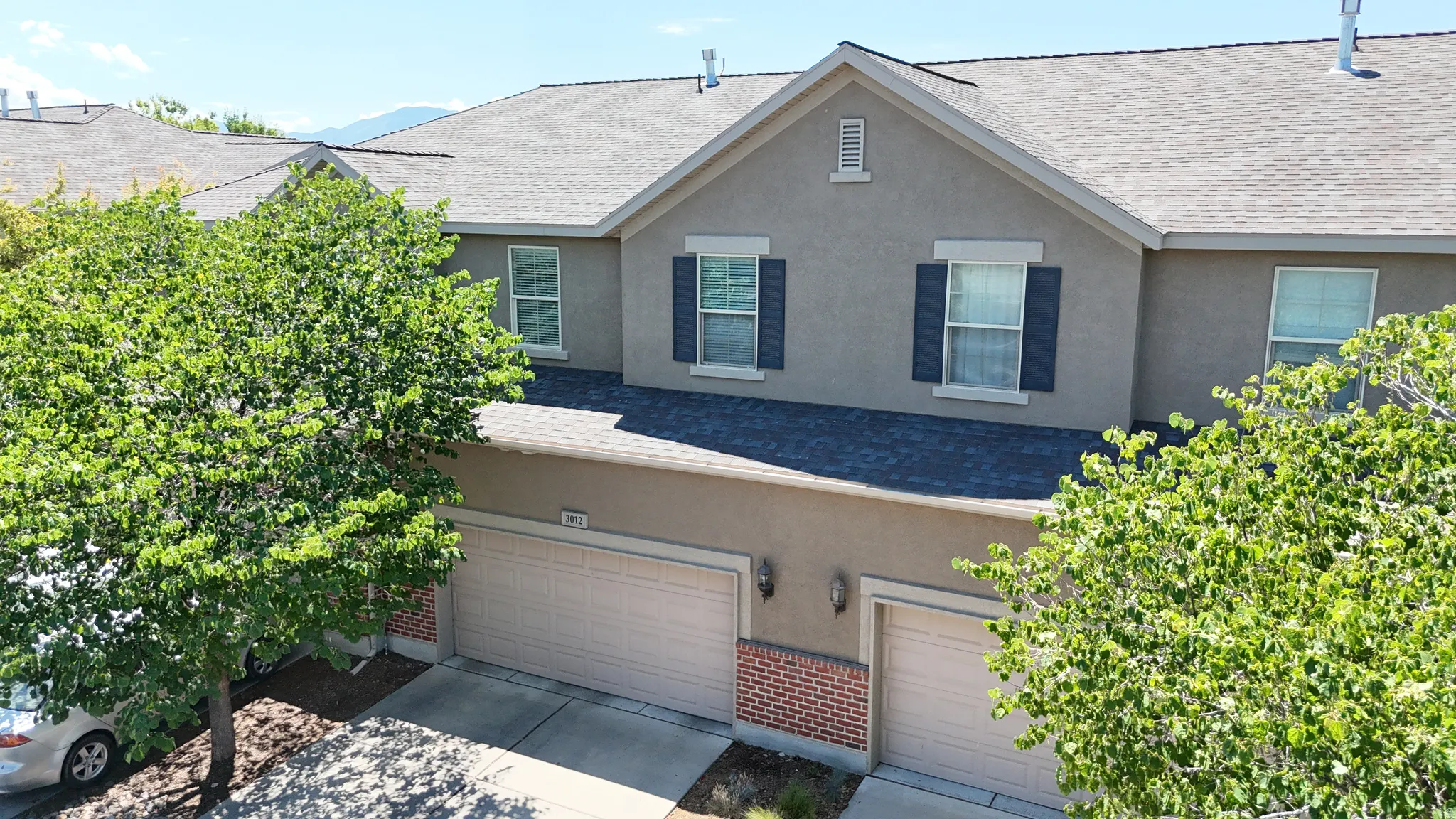 Stucco exterior with brick accents, featuring two attached garages, a pitched shingle roof, and multiple windows with dark shutters