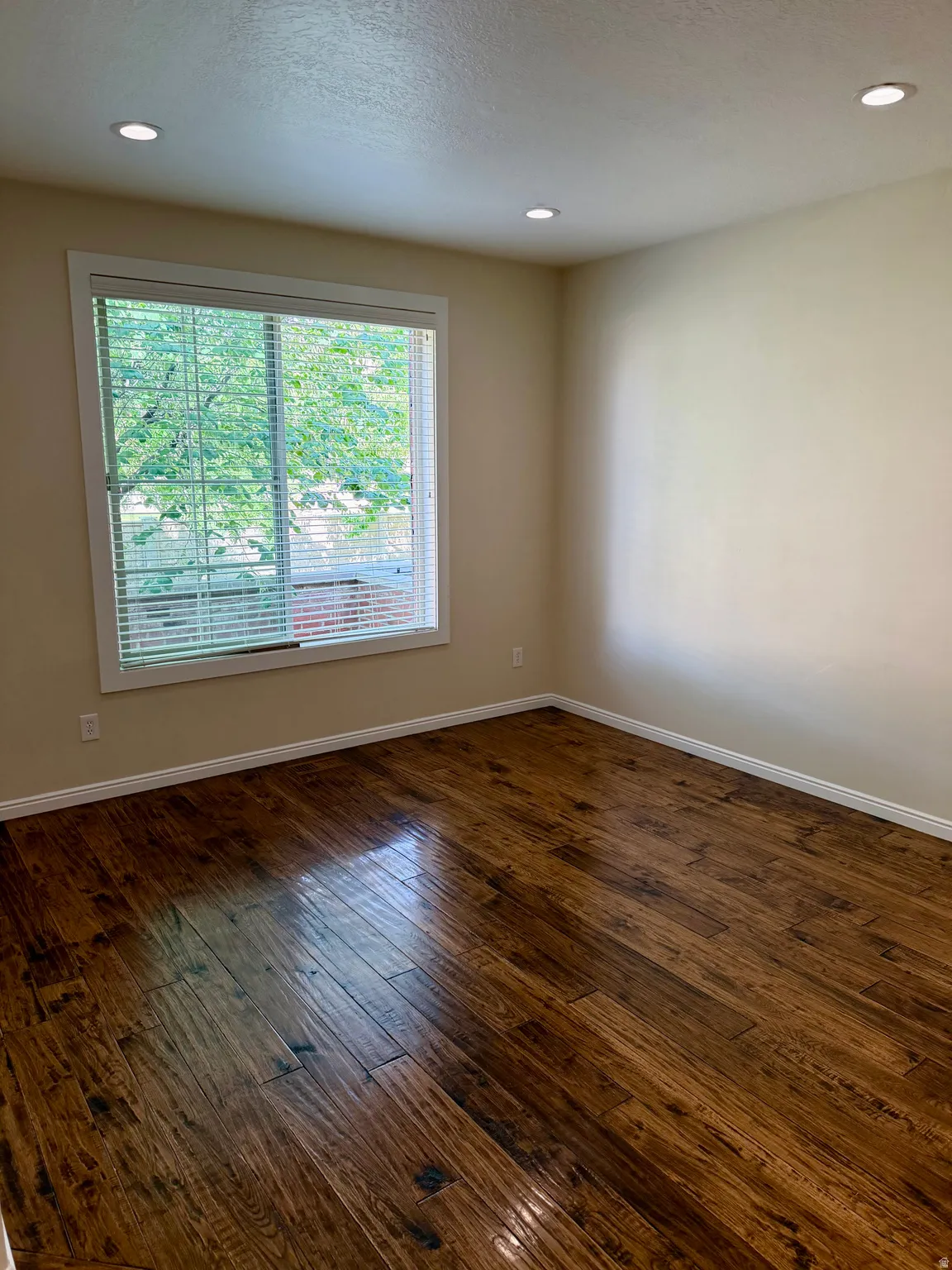 Spacious room featuring rich wood-finish flooring, recessed lighting, neutral wall tones, and a large window with horizontal blinds