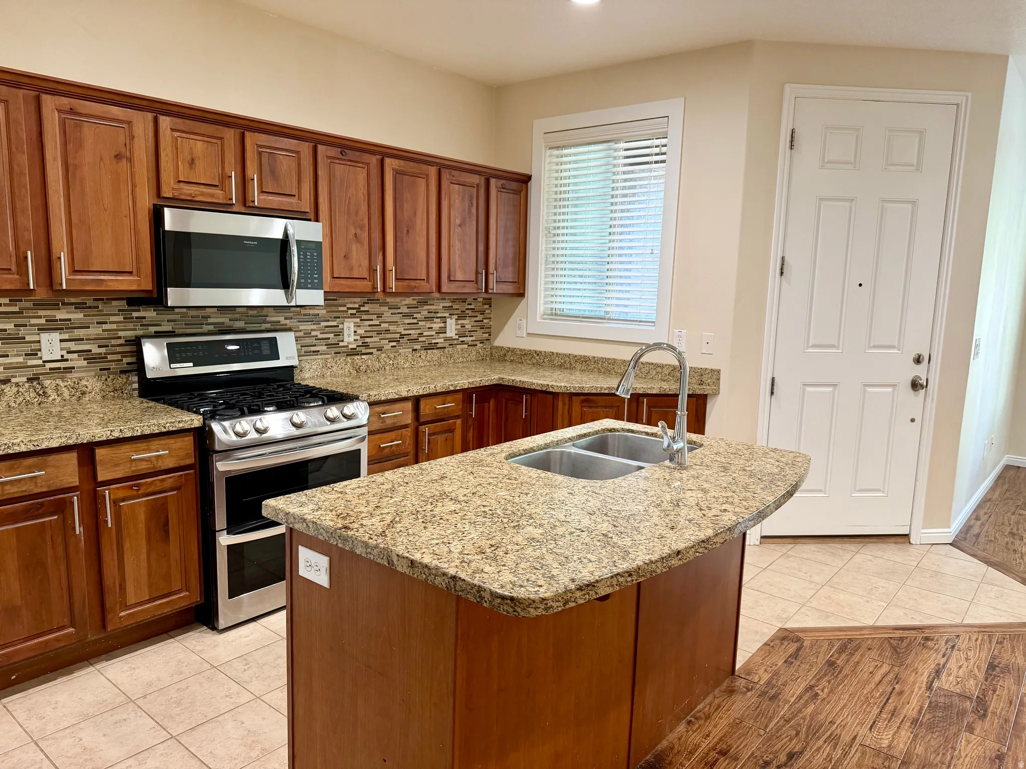 Kitchen featuring granite countertops, wood cabinetry, stainless steel appliances, a tile backsplash, and a central island with a double basin sink