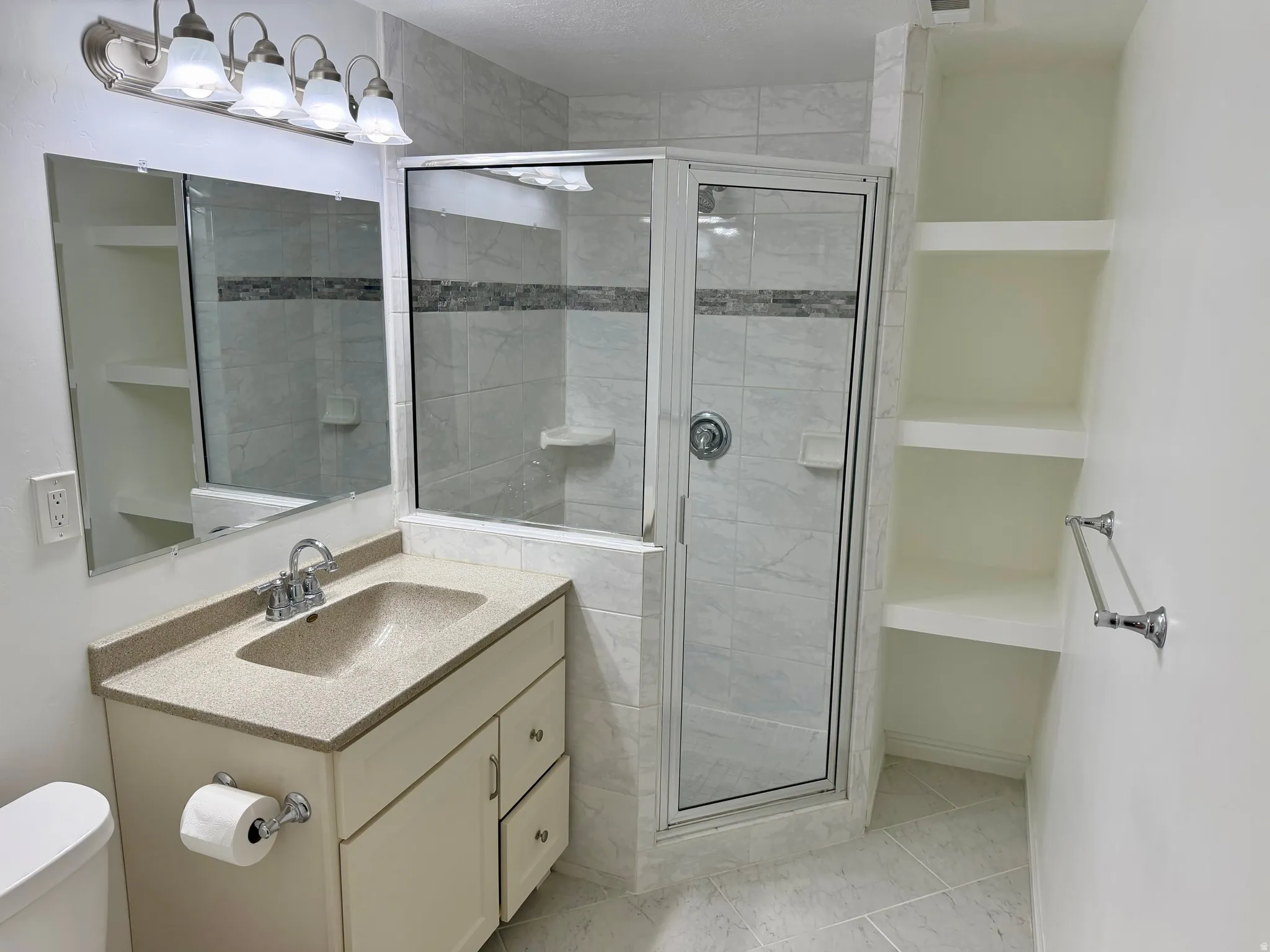 Bathroom featuring a glass-enclosed shower with decorative tile accent, a single vanity with an integrated sink and light-toned countertop, a large wall-mounted mirror, and built-in shelving