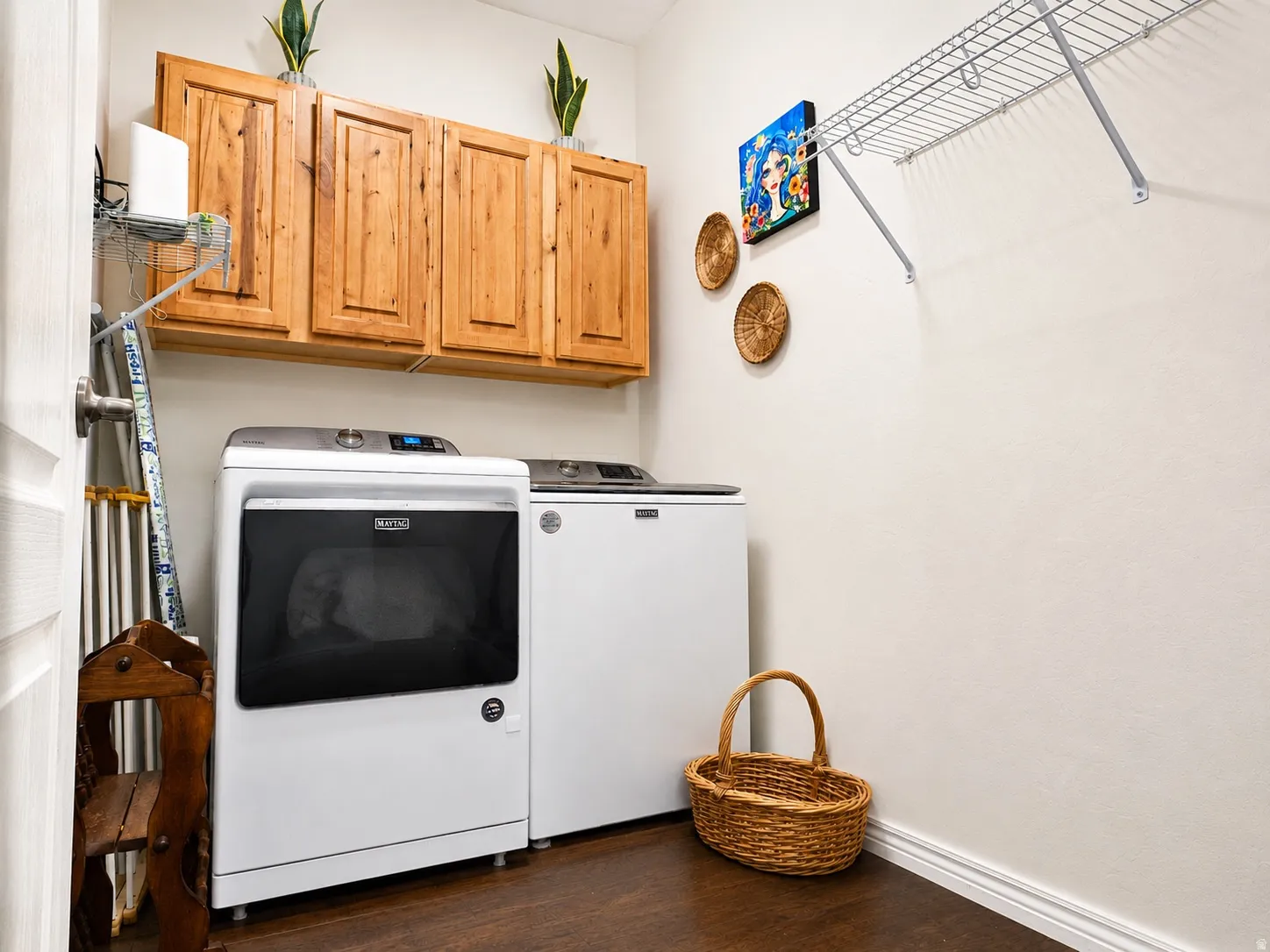 Laundry area featuring wood-finish flooring, light-toned walls, built-in upper cabinetry with wood panel doors, and a mounted wire shelf