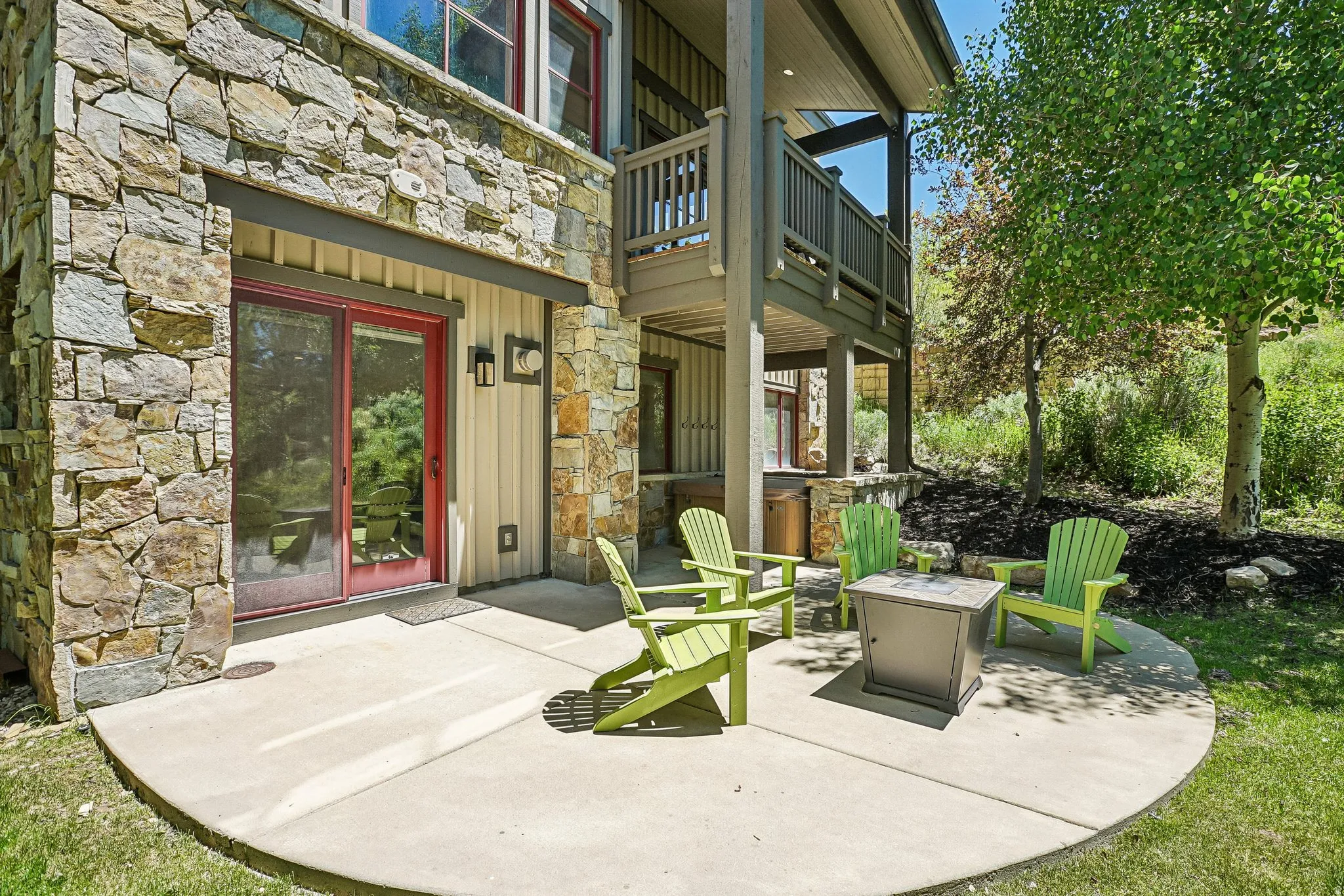 Lower-level patio with a stone and siding exterior, featuring a sliding glass door with a red frame, and an upper-level balcony