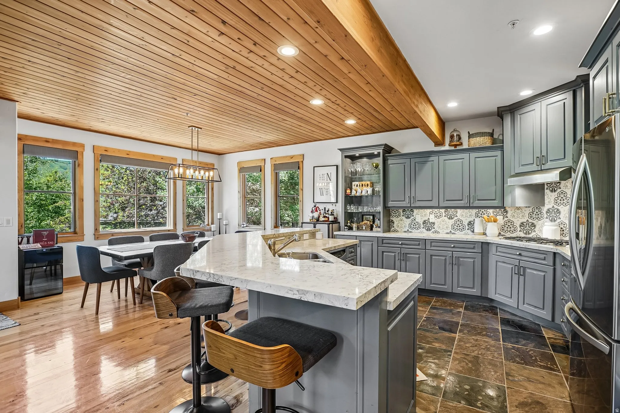 Open-concept kitchen and dining area featuring a wood-panel ceiling with exposed beam, recessed lighting, and a linear chandelier