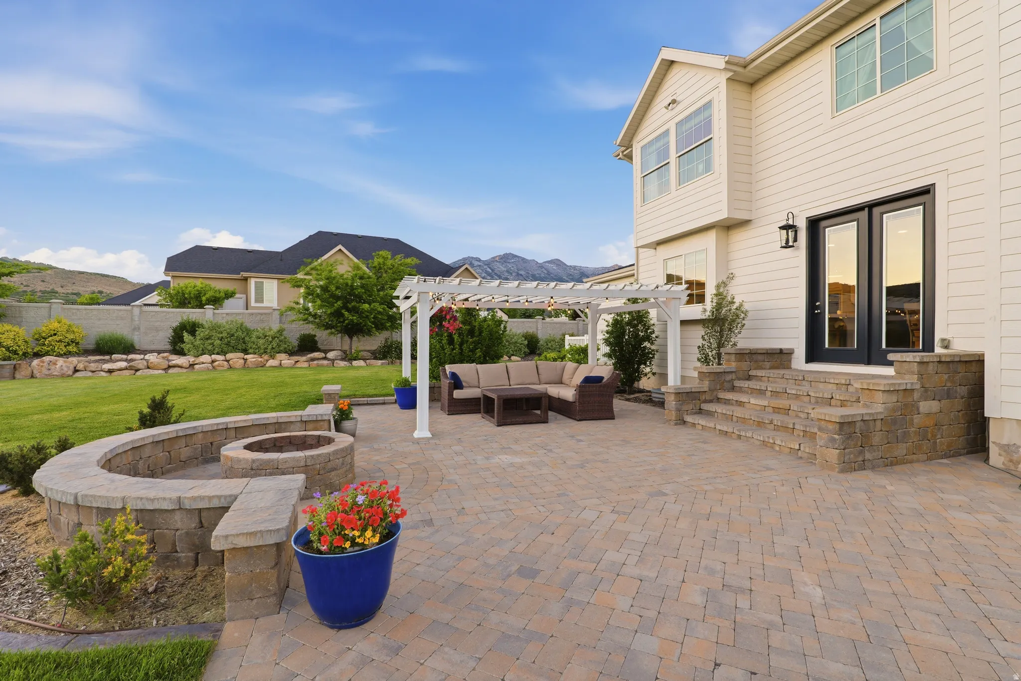 Paver patio with a white pergola, built-in fire pit with stone seating, stone steps, and dark-framed double doors