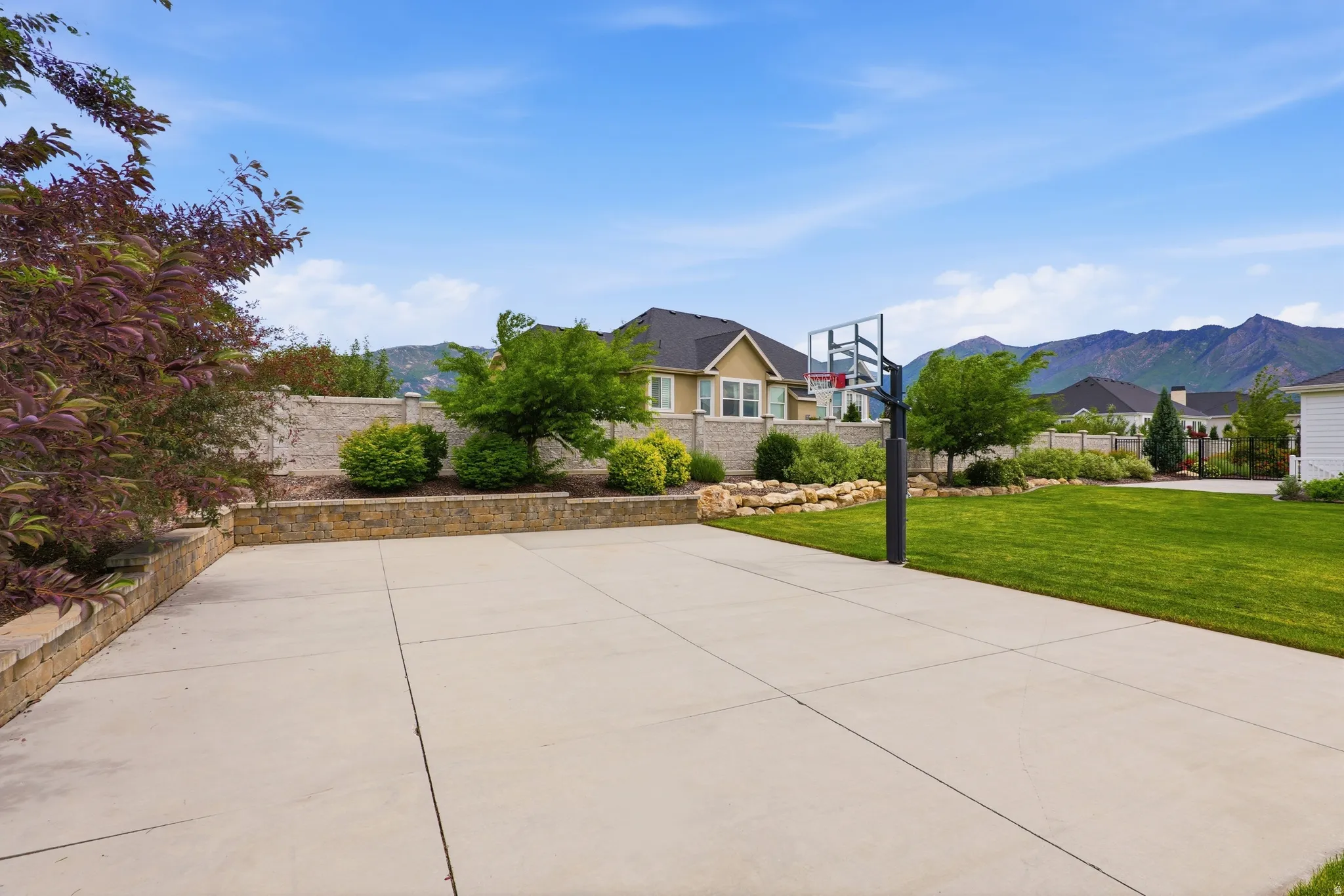Expansive concrete pad with a basketball hoop, bordered by a stone retaining wall and mature landscaping