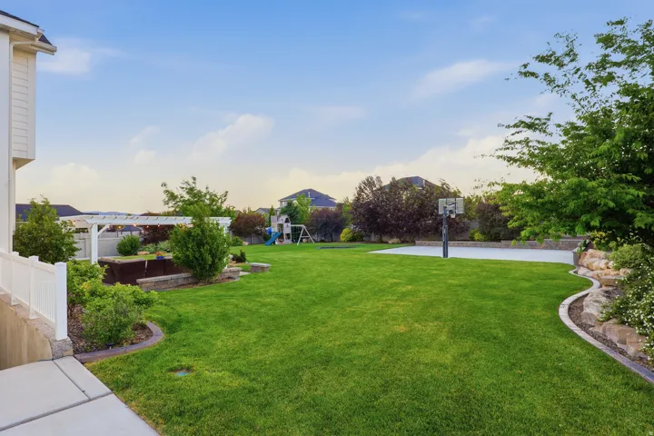 Expansive backyard featuring a large green lawn, a white pergola, a dedicated sports court, mature trees, and layered landscaping with stone retaining walls