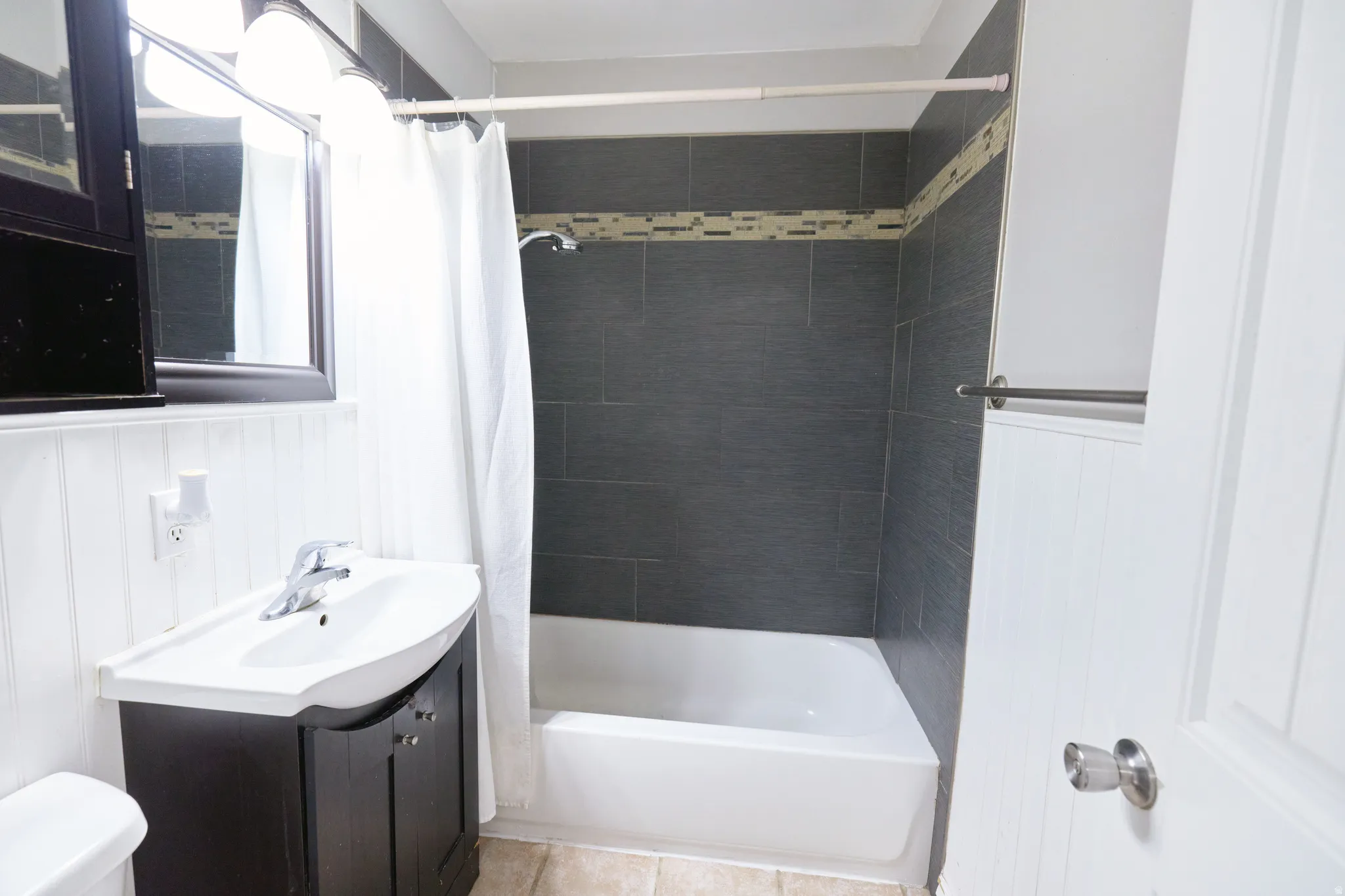 Bathroom featuring a dark wood vanity with white basin sink, chrome faucet, white beadboard wall paneling, and a shower-over-tub with dark gray subway tile surround and decorative accent band