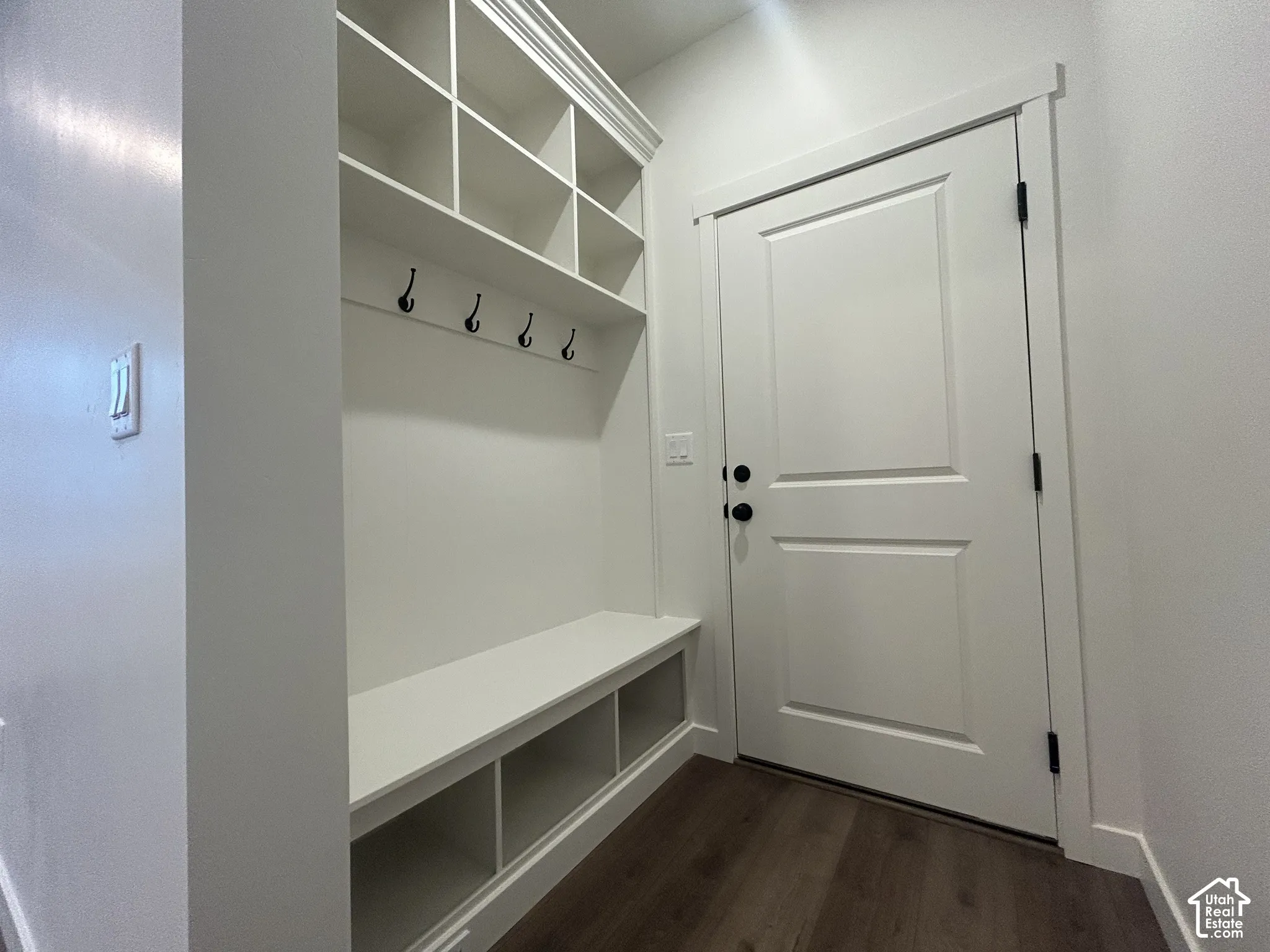 Built-in mudroom cabinetry featuring upper cubbies, four coat hooks, and a bench with lower storage compartments, complemented by wood-finish flooring and a paneled entry door with matte black hardware