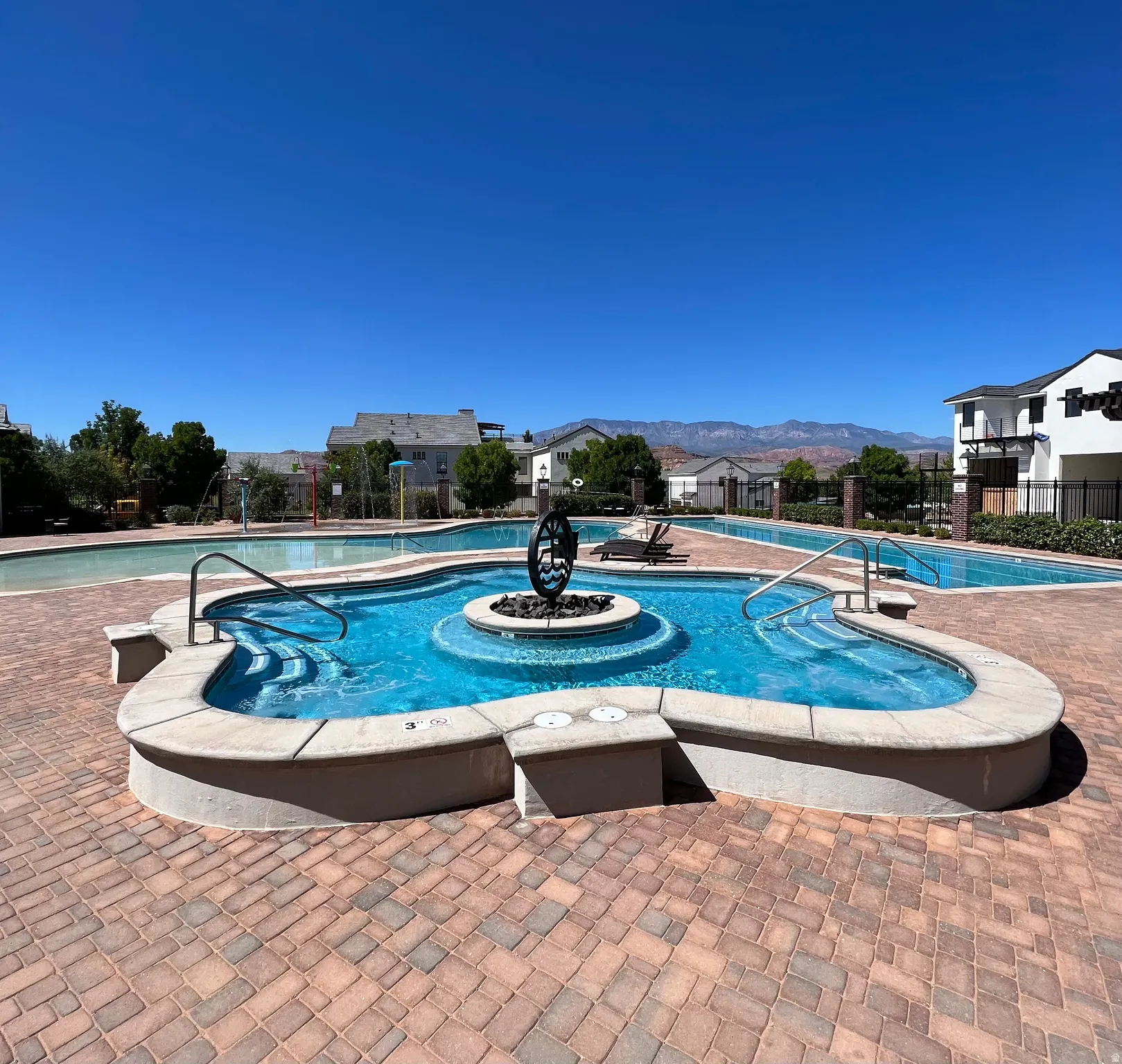 Resort-style pool area featuring a curved spa with a central decorative element, a large swimming pool, and a textured paver deck