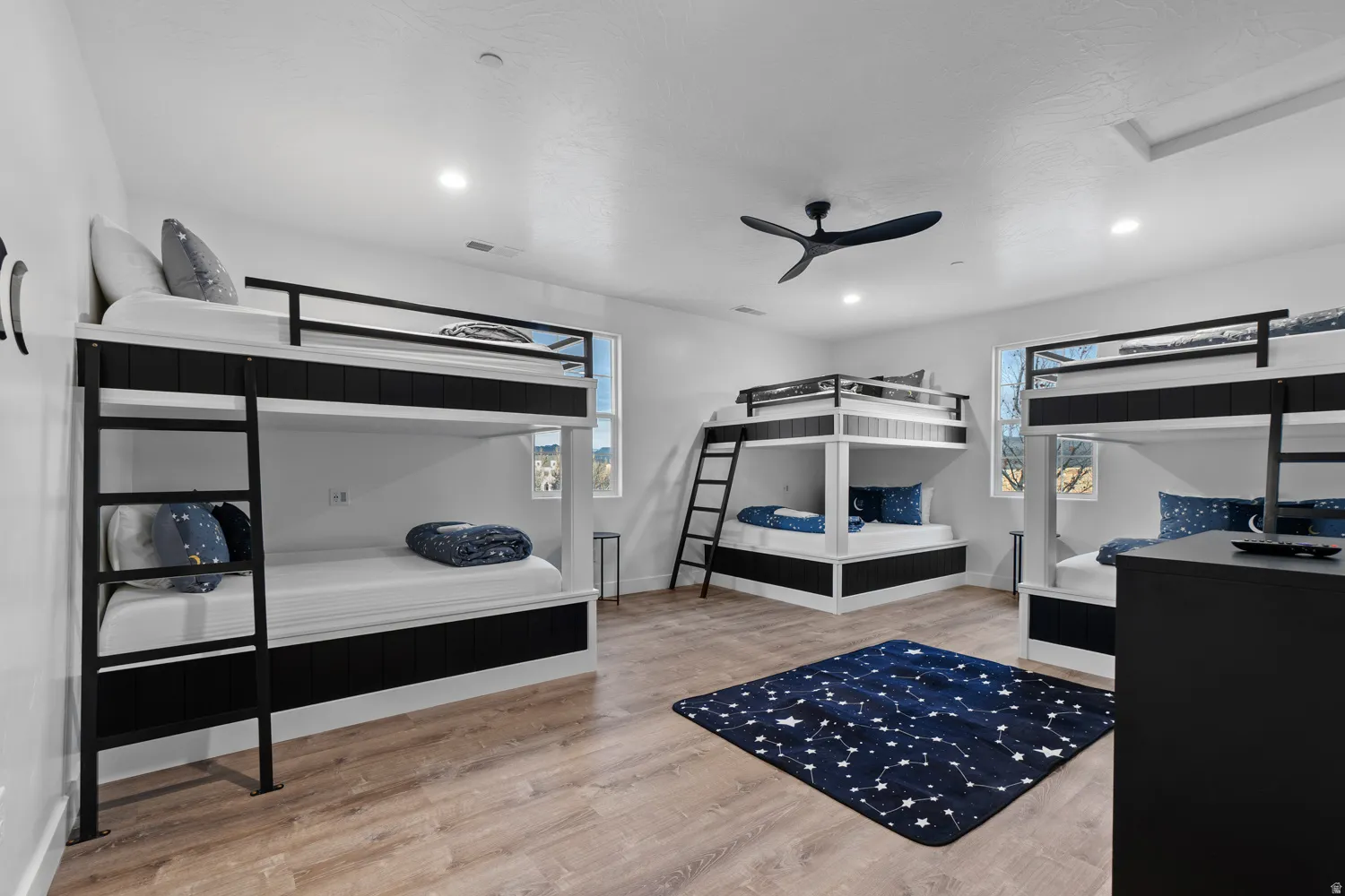 Spacious room featuring multiple built-in bunk beds with black and white frames, light wood-finish flooring, recessed lighting, and a contemporary ceiling fan
