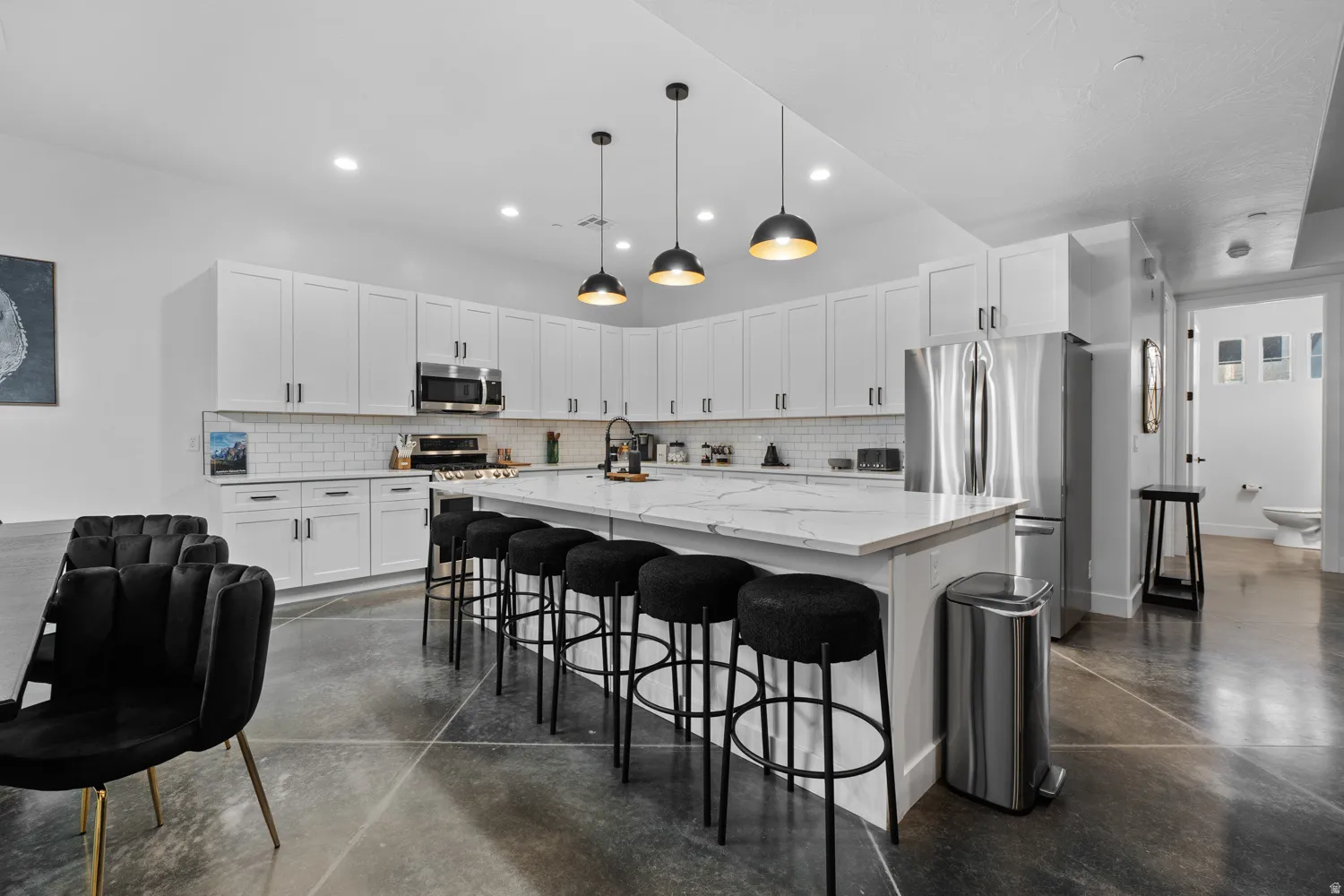 Open-concept kitchen featuring an oversized island with seating, stainless steel appliances, white shaker cabinetry, subway tile backsplash, and concrete flooring