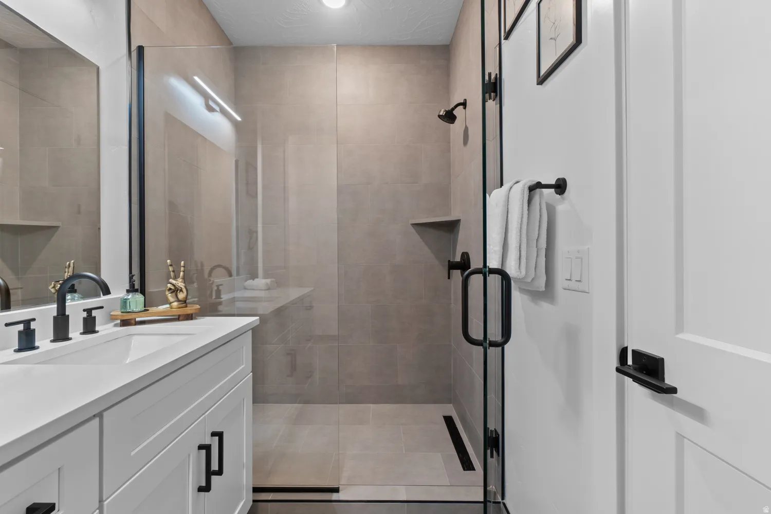 Bathroom featuring a frameless glass shower enclosure, a single vanity with a white countertop and rectangular undermount sink, matte black fixtures, and light-toned tile flooring and shower surround