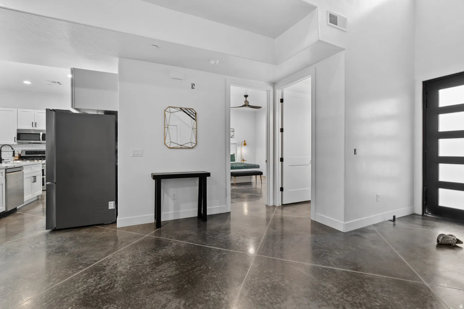 Polished concrete flooring spanning an open-concept interior featuring a contemporary black entry door with frosted glass panels, a kitchen area with white cabinetry and stainless steel appliances, and a recessed lighting design