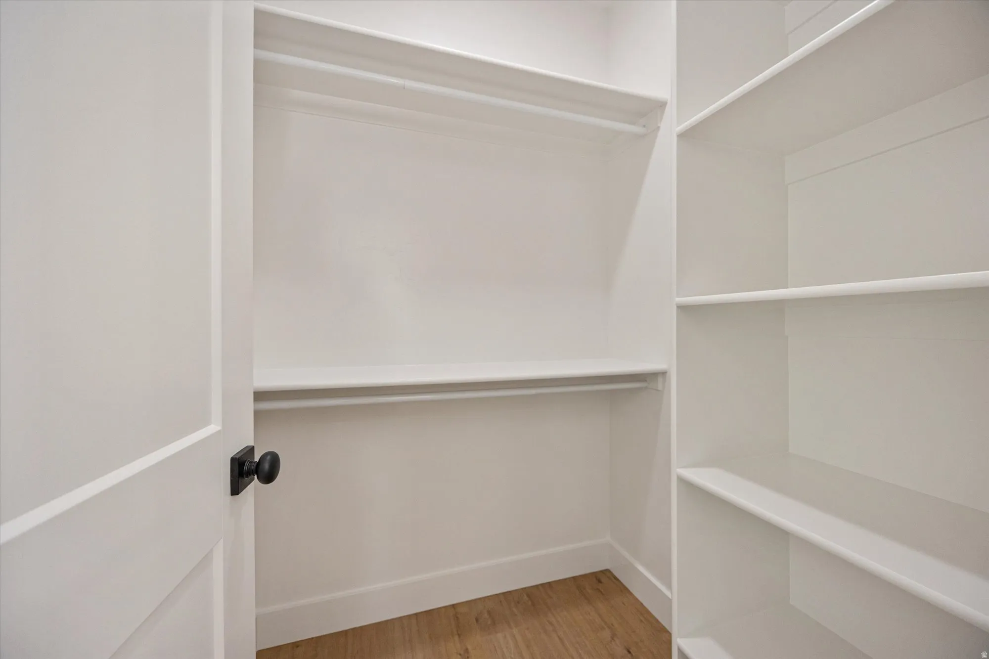 White walk-in closet featuring built-in shelving, two hanging rods, a white paneled door with matte black hardware, and wood-finish flooring
