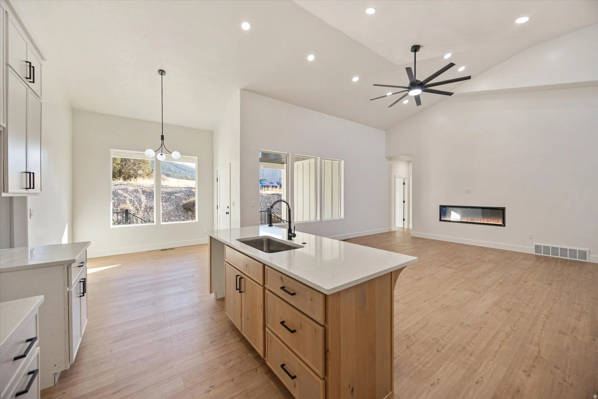 Open-concept living space featuring a central kitchen island with a white countertop and natural wood-finish cabinetry, wood-finish flooring, and a modern linear fireplace